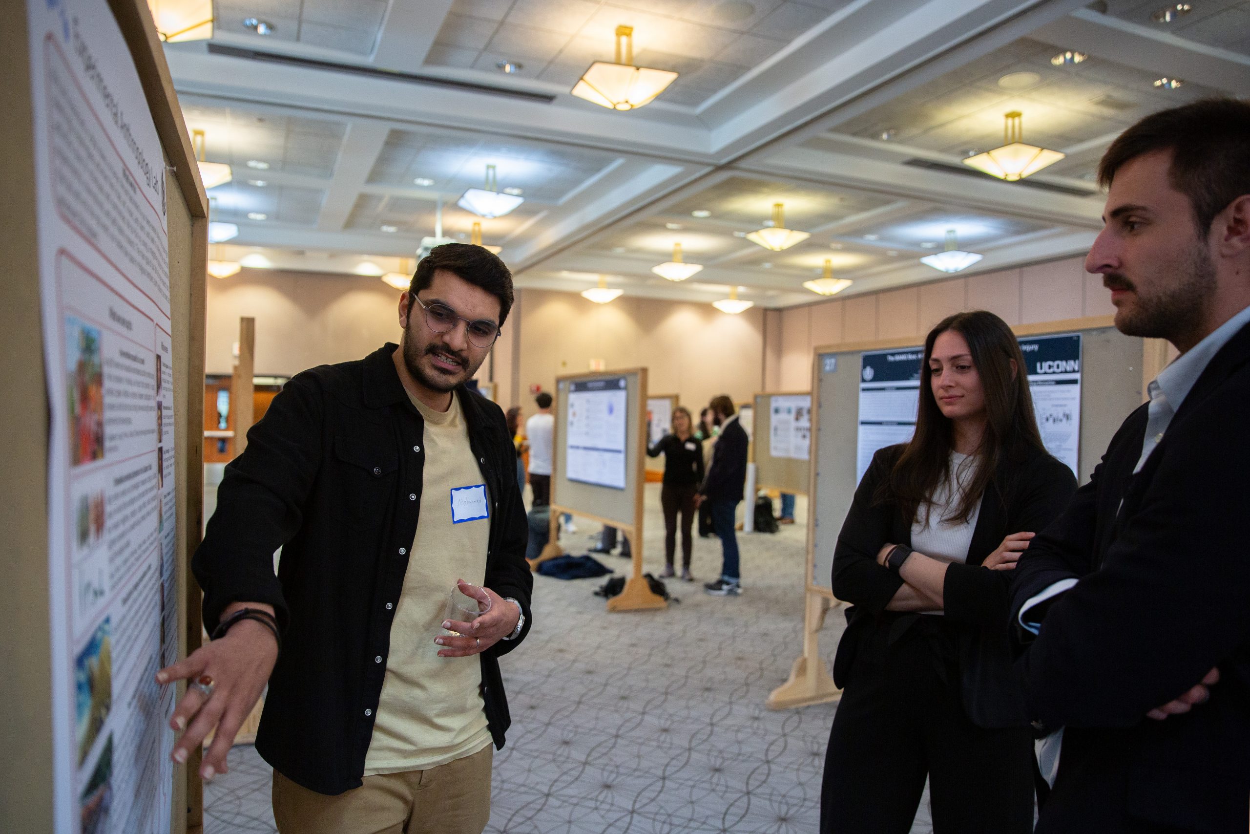 A graduate student presents a research poster to two attendees, using hand gestures to explain their work